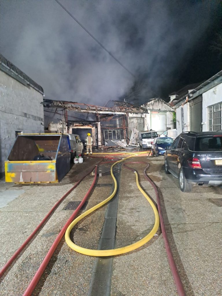 Fordingbridge incident photo showing the metal frame of an industrial unit in the background. There is smoke emanating from the building, and the roof and walls are almost completely burnt away. In the foreground there are firefighting hoses laid out on the ground, and a firefighter is assessing the building. 