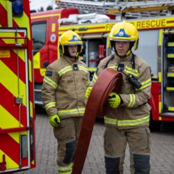 two firefighters with an engine in the back ground. The firefighters are coming around the side of another fire engine of which you can see a small portion of the back. One firefighter is carrying a hose