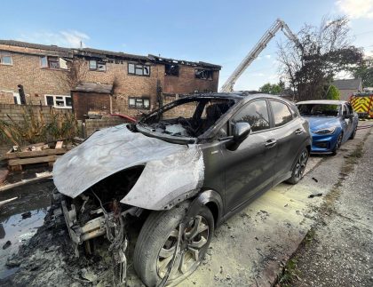 Two partially burnt cars sits in the foreground, having been extinguished by firefighters. In the background is a row of terrace houses with fire damage to the interior and exterior of the property. A fire engine can be seen parked behind the burnt vehicles.