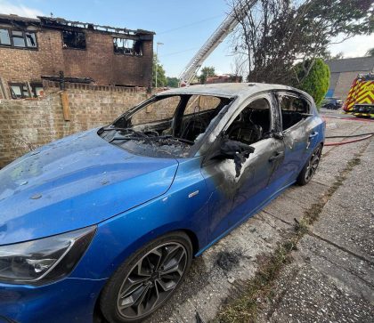 A partially burnt car sits in the foreground, having been extinguished by firefighters. In the background is a row of terrace houses with fire damage to the interior and exterior of the property. A fire engine can be seen parked behind the burnt vehicle.